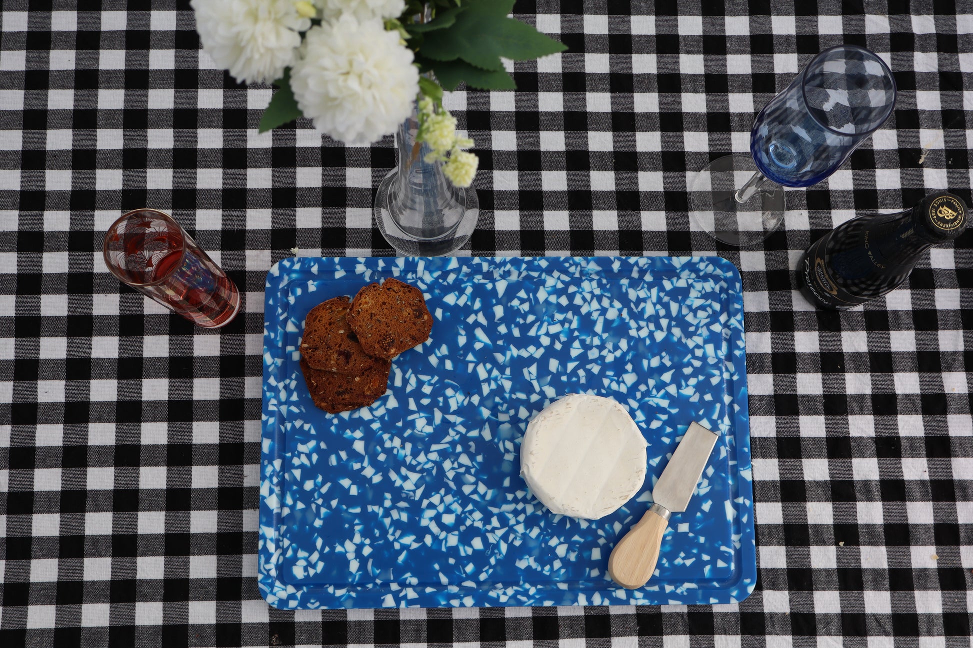Blue speckled cutting board with cheese, crackers, and a knife on a black and white checkered tablecloth.