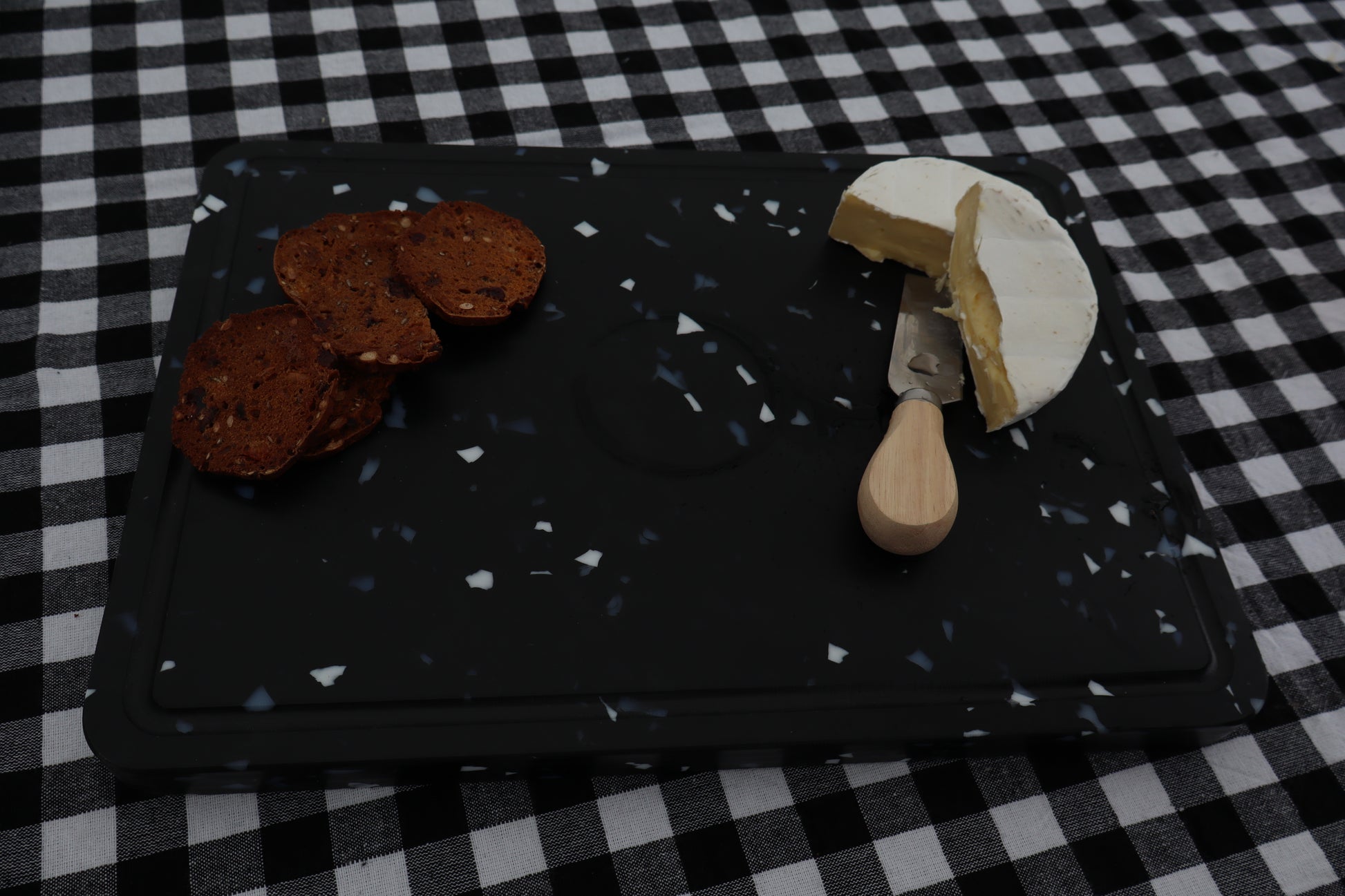 Black chopping board with crackers and cheese on a black and white checkered tablecloth.