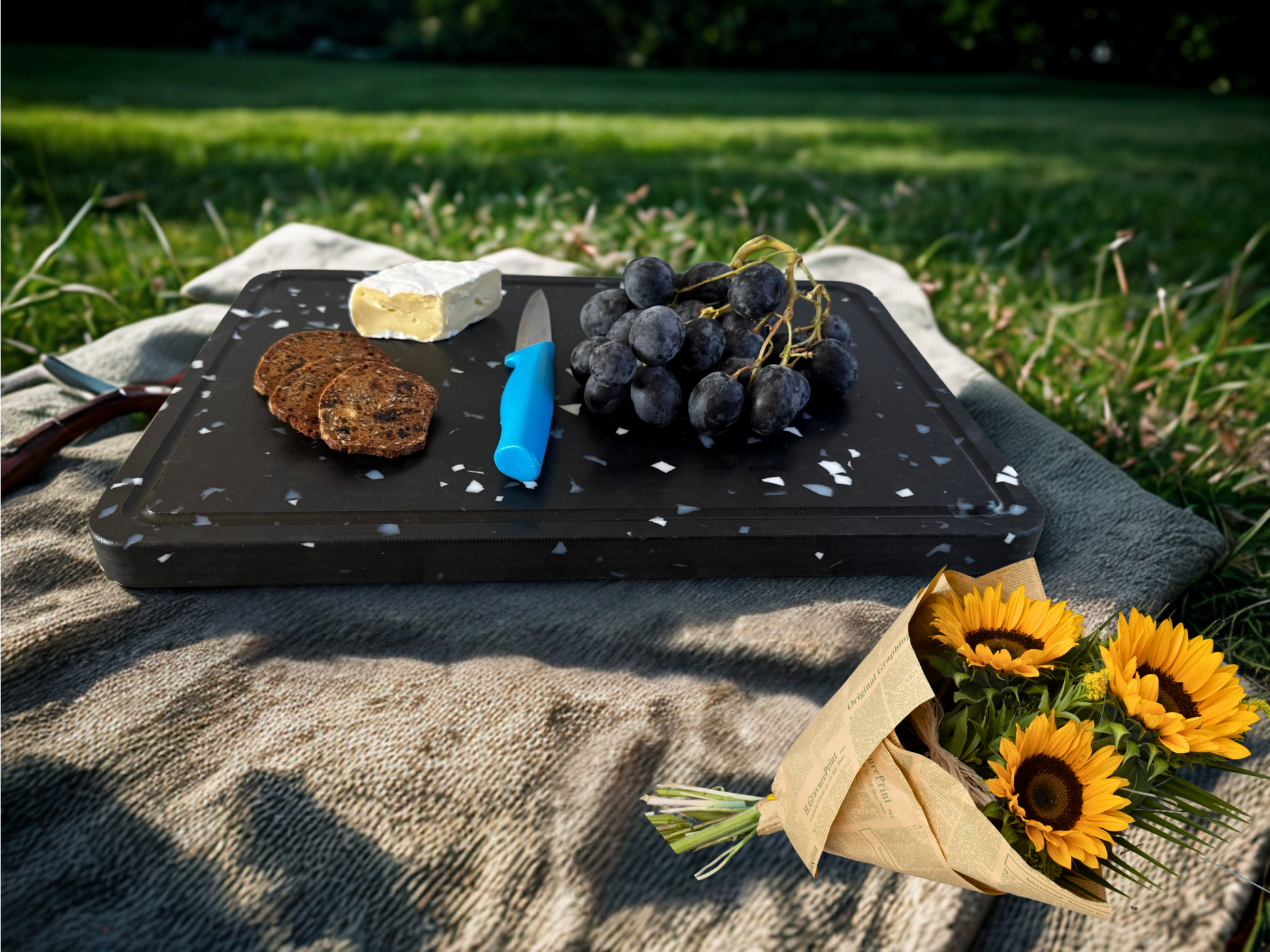 Black cutting board with grapes, bread, and a knife on a picnic blanket outdoors.